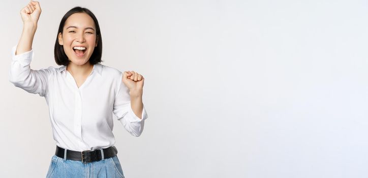 Image of happy lucky asian woman hooray gesture, winning and celebrating, triumphing, raising hadns up and laughing, standing over white background.