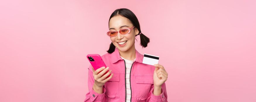 Online shopping. Smiling asian girl shopper, holding smartphone and credit card, paying in mobile app, standing over pink background.