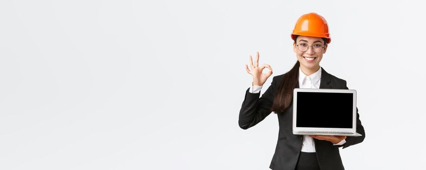 Smiling professional female architect, construction manager at factory showing graph, positive diagram, make okay gesture in approval and holding laptop facing screen, wear safety helmet.