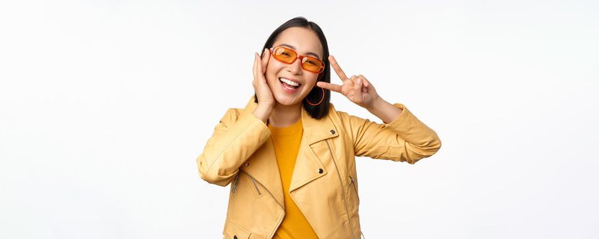 Portrait of stylish asian modern girl, wearing sunglasses and yellow jacket, showing peace, v-sign gesture, standing over white background, happy smiling face.