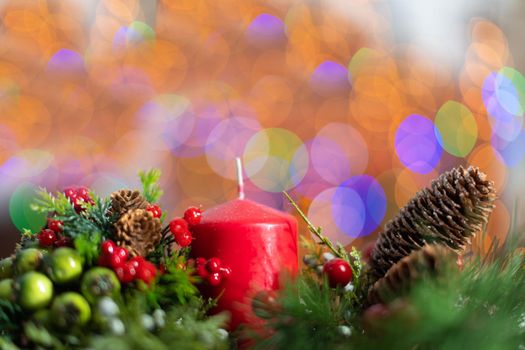 A close up shot shows a red candle sticking out of a Christmas wreath. In the distance a blurred background created from the bokeh of the glowing Christmas tree lights