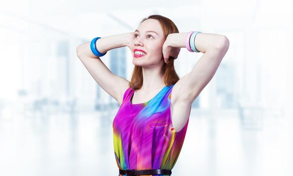 Redhead beautiful girl closed her ears with hands. Joyful lady with charming smile in bright colorful dress and bracelets on blurred office background. Portrait of happy young woman looking up aside.