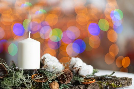 In the close up frame you can see a white candle sticking out of a Christmas wreath. In the distance a blurred background created from the bokeh of the glowing Christmas tree lights