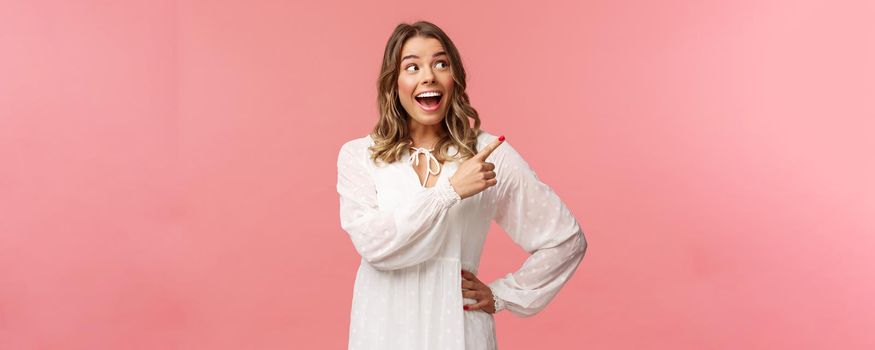 Enthusiastic, happy attractive blond woman in white cute dress, open mouth excited and cheerful looking, pointing upper right corner as best prices discounts ever, stand pink background.