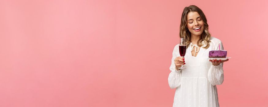 Holidays, spring and party concept. Portrait of tender, elegant young blond woman holding glass of wine and cake with lit candle, smiling pleased, celebrating birthday, pink background.