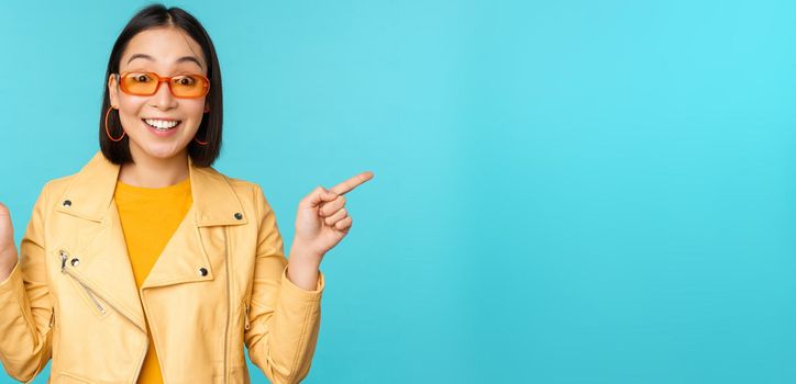Happy stylish chinese girl in sunglasses, points fingers sideways, invites to check out, see store discount, shows left and right, stands over blue background.