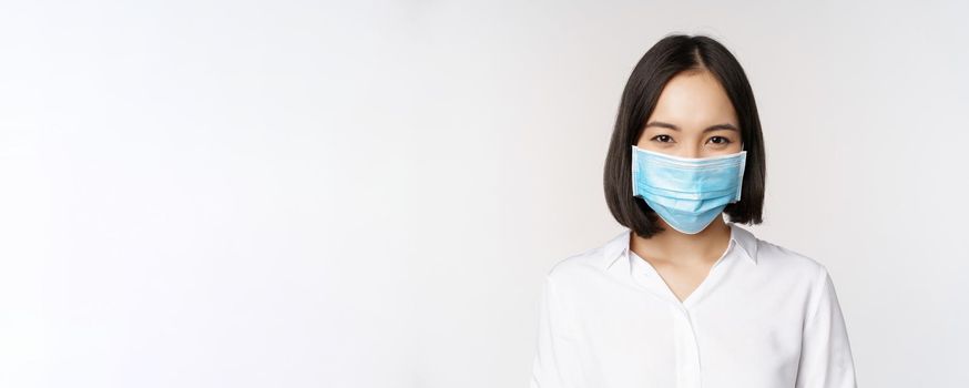 Covid and healthcare concept. Close up portrait of asian woman, office lady in face mask, smiling, using protection from coronavirus during pandemic, white background.