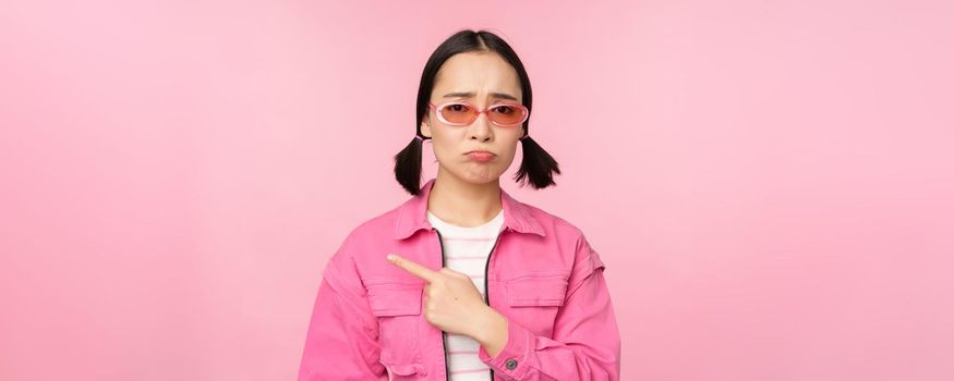 Potrait of cute korean girl in sunglasses, pointing left and looking disappointed, sulking upset, standing over pink background.