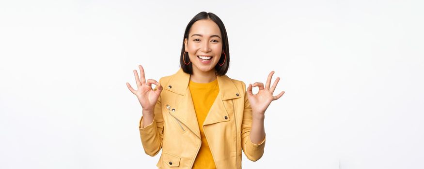 Happy beautiful asian girl shows okay, ok recommending sign, pleased by smth, praising quality, standing over white background. Copy space