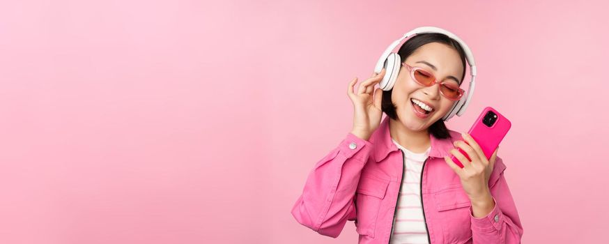 Stylish asian girl dancing with smartphone, listening music in headphones on mobile phone app, smiling and laughing, posing against pink background.