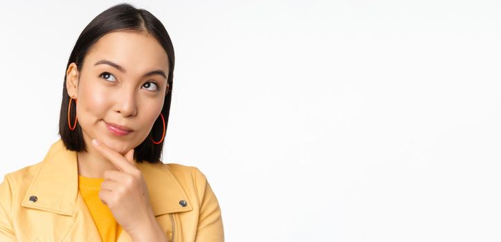 Close up of asian woman thinking, looking up thoughtful, pondering smth with pleased smiling face, standing over white background.