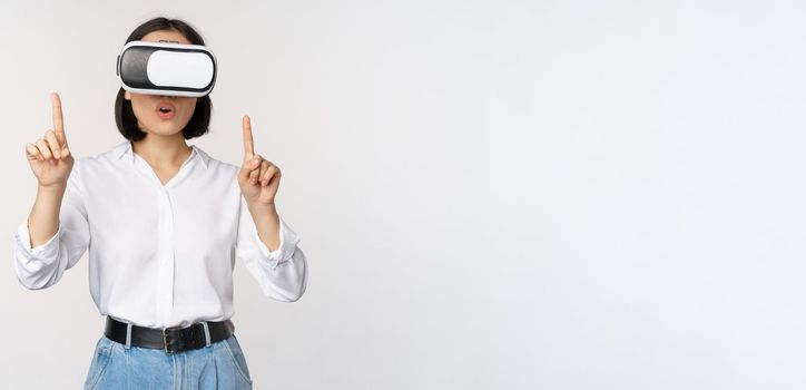 Image of asian woman in vr glasses pointing fingers up, looking amazed and surprised. Girl using virtual reality headset, concept of future and communication, white background.