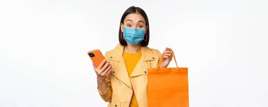 Image of stylish asian girl shopper, holding smartphone and shopping bag without store logo, wearing medical face mask from covid-19, white background.