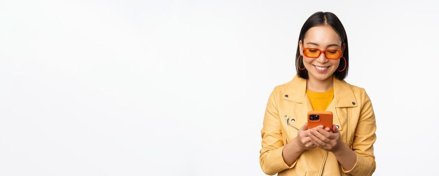 Modern asian girl in sunglasses using her mobile phone, smiling and looking happy, posing against white background. Copy space