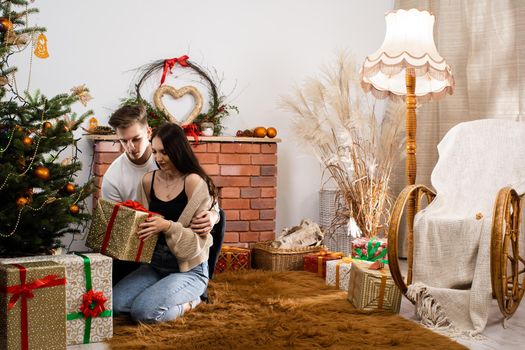 Two lovers are sitting by the Christmas tree on Christmas. Decorated Christmas tree and fireplace in the living room at home.