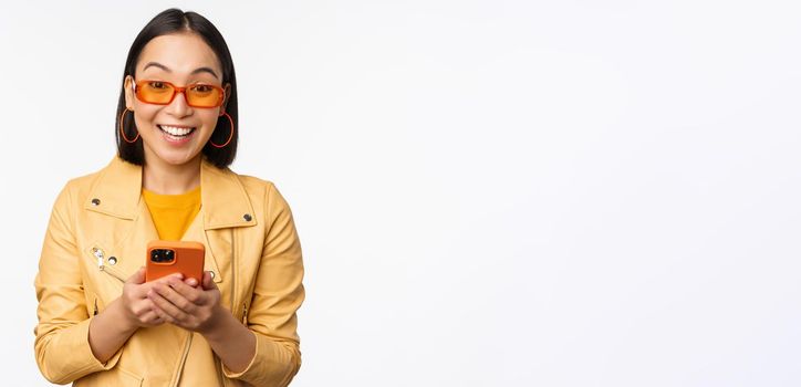 Modern asian girl in sunglasses using her mobile phone, smiling and looking happy, posing against white background. Copy space