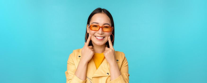 Close up portrait of asian young woman in sunglasses, smiling and looking romantic, standing happy over blue background. Copy space