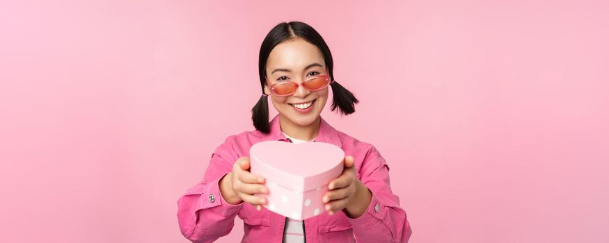 Cute asian girl giving you gift in heart shaped box, kissing and smiling, concept of holiday and celebration, standing over pink background.