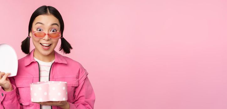 Cute asian girl giving you gift in heart shaped box, kissing and smiling, concept of holiday and celebration, standing over pink background.