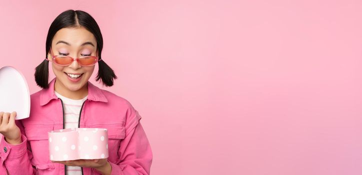 Beautiful asian girl opens up heart shaped gift box, looking happy, standing in sunglasses, smiling surprised at camera, pink background.