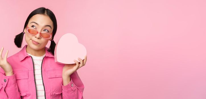 Image of stylish asian girlfriend in sunglasses, guessing whats inside gift box, heart-shaped present, standing over pink background.