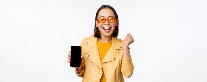 Happy asian girl in sunglasses, showing mobile phone screen, smartphone interface, laughing and smiling, celebrating, standing over white background.