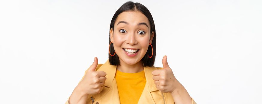 Smiling asian girl showing thumbs up, looking pleased, approve smth, standing over white background. Copy space