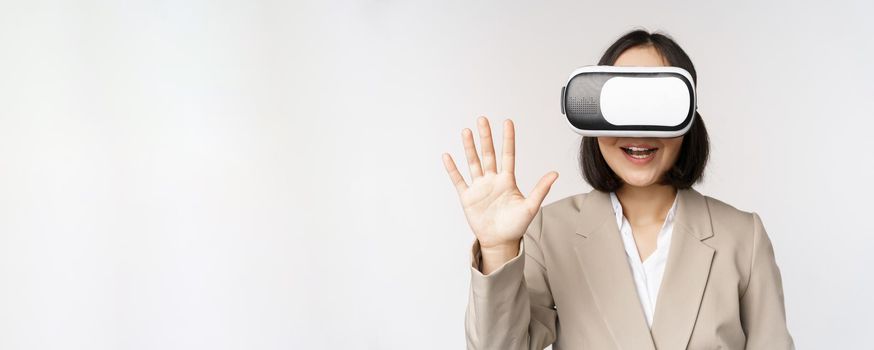 Meeting in vr chat. Asian businesswoman in virtual reality glasses, waving hand and saying hello, greeting someone, standing over white background.