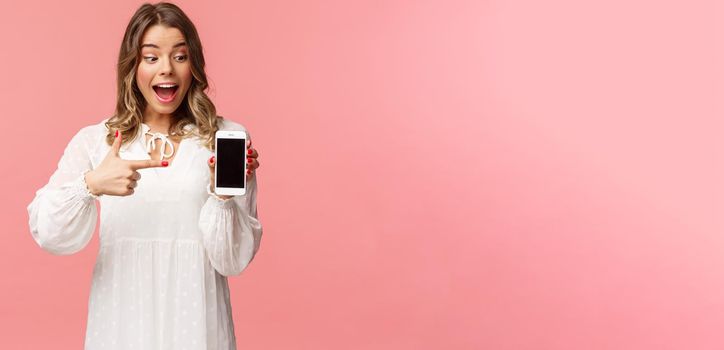 Portrait of excited and amazed, happy blond female in white dress, pointing finger at mobile phone screen, looking at smartphone with pleased and surprised smile, stand pink background.