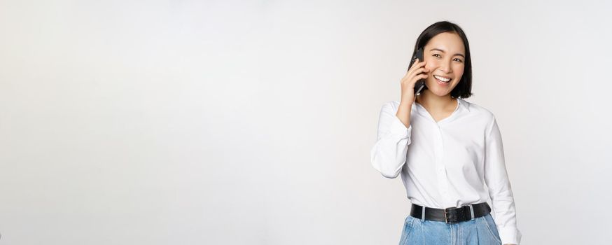 Friendly smiling asian woman talking on phone, girl on call, holding smartphone and laughing, speaking, standing over white background.