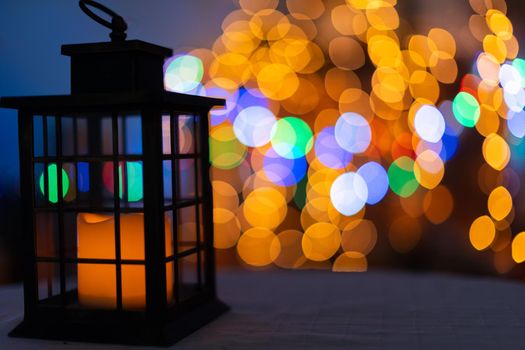 In the night frame a rectangular old hand lantern with a glowing candle in the center. In the distance a blurred background created from the bokeh of glowing Christmas tree lights