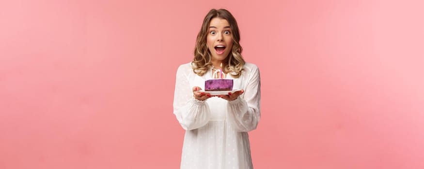 Holidays, spring and party concept. Happy cheerful good-looking blond woman celebrating birthday, holding piece cake with lit candle, making wish, look amused standing pink background.