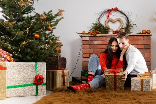 The bride and groom, sitting by the Christmas tree, communicate their feelings in body language. Lovers flirt in a decorated living room at home. December Christian holidays.