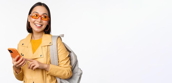 Stylish young asian woman tourist, traveller with backpack and smartphone smiling at camera, posing against white background. Copy space