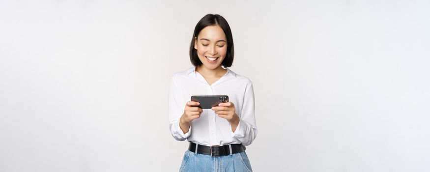 Young korean woman, asian girl playing mobile video game on smartphone, looking at horizontal phone screen, standing over white background.