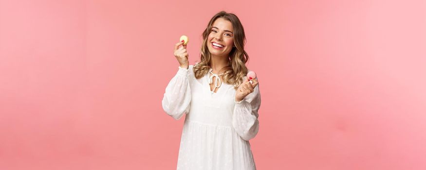 Holidays, spring and party concept. Portrait of cute romantic blond girl in white dress, licking lips as tempting to eat delicious dessert, holding two macarons and look pleased, pink background.