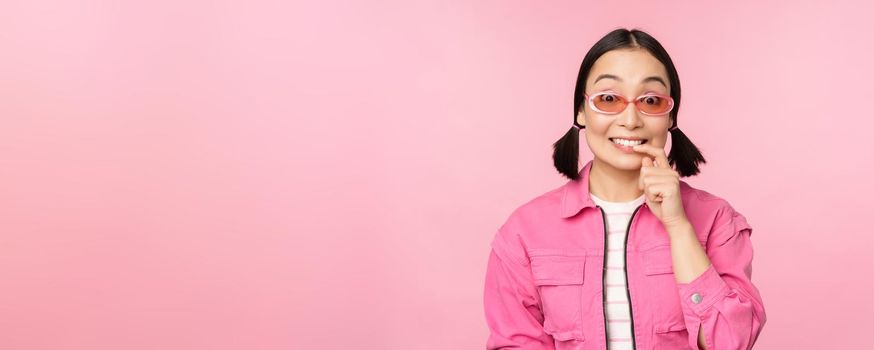 Stylish asian girl in sunglasses, looks thoughtful or intrigued, wants to try smth out, stands over pink background, pondering.
