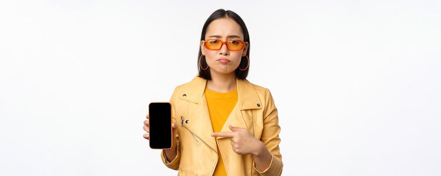 Portrait of sad asian woman in sunglasses, pointing finger at mobile phone app interface, showing smartphone application, standing over white background.