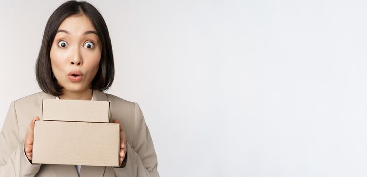 Portrait of asian saleswoman looking surprised, holding boxes, delivery goods, standing amazed in suit against white background.