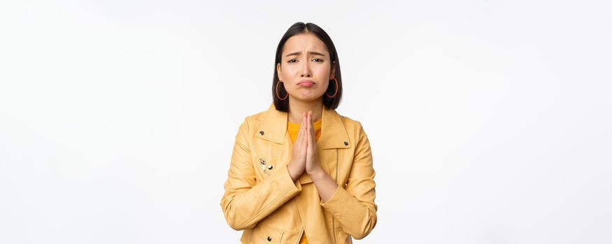 Image of hopeless asian woman begging, pleading, say sorry, looking distressed and miserable, standing over white background.