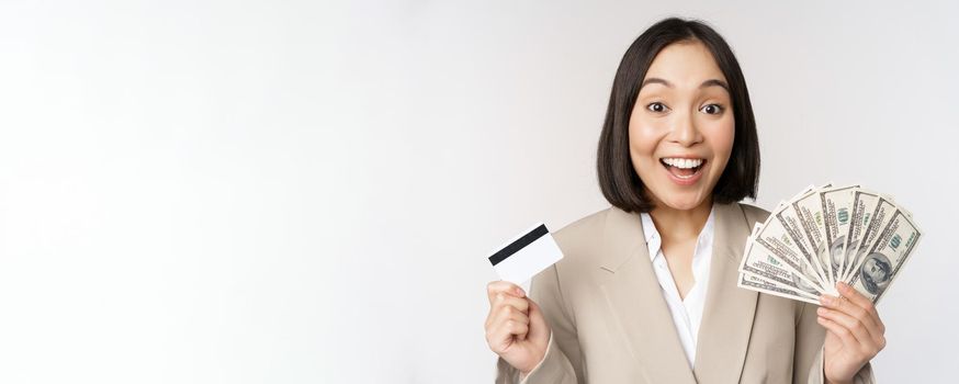 Enthusiastic young asian businesswoman, showing money dollars and credit card, standing over white background.