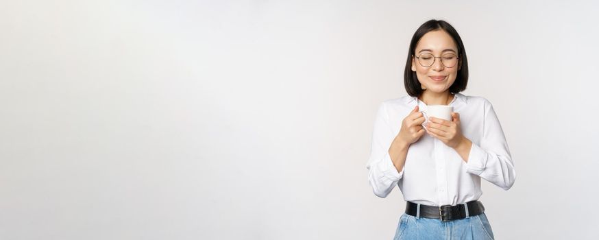 Image of smiling korean woman sniffing, smell coffee in mug, enjoying delicious drink, standing over white background.