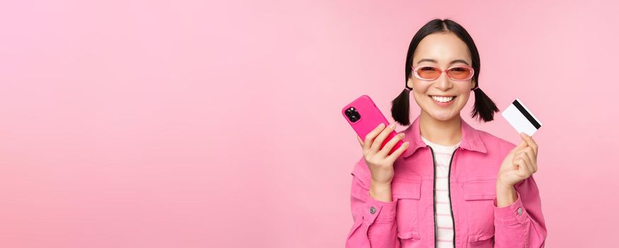Beautiful korean woman holding smartphone, credit card, smiling at camera, buying online, shopping with mobile phone, standing over pink background.