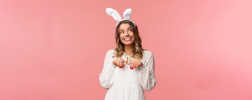 Holidays, spring and party concept. Cute romantic young blond girl imitating bunny, wear white dress lovely rabbit ears, make hand-paws near chest and look dreamy up with daydreaming smile.