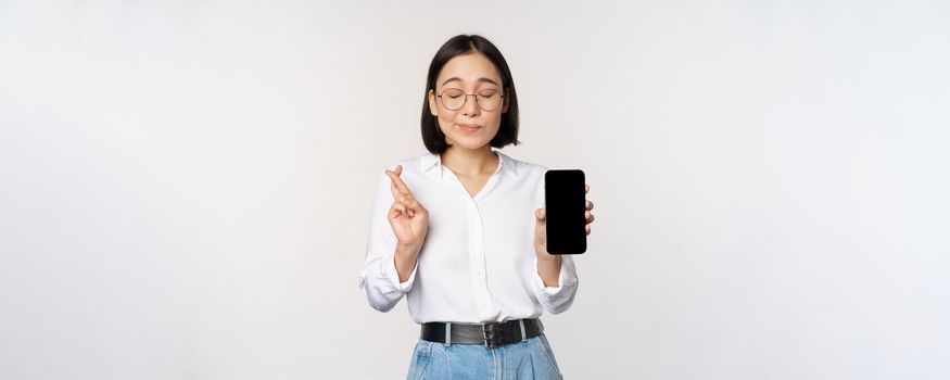 Hopeful young asian woman, showing mobile phone screen, app interface and fingers crossed, hoping for smth, making wish or paying, standing over white background.