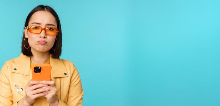 Image of sad asian girl in sunglasses, looking disappointed, holding mobile phone, standing over blue background.