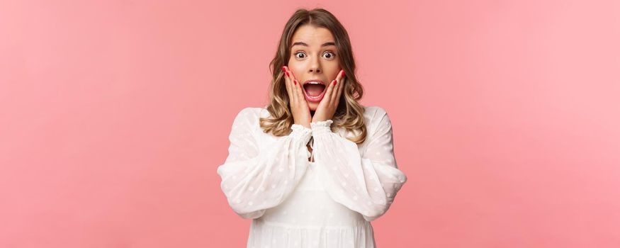 Happy and surprised blond girl hear awesome news, gasping speechless staring camera with hands on cheeks, being impressed and stunned over great event, standing pink background.