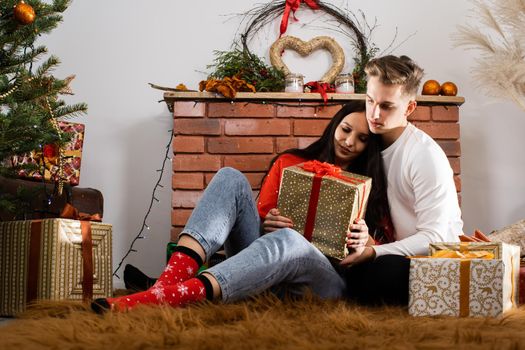The bride and groom, sitting by the Christmas tree, communicate their feelings in body language. Lovers flirt in a decorated living room at home. December Christian holidays.