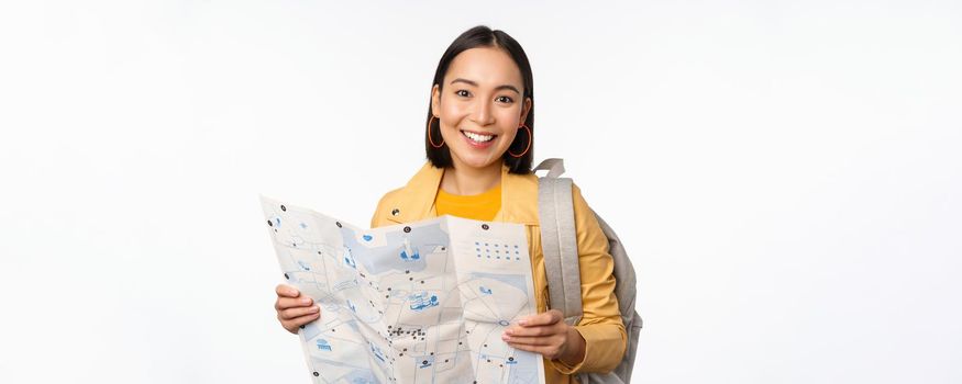 Tourism and backpacking. Smiling asian girl holding map and backpack, tourist searching way, travelling, standing over white background.