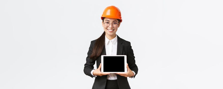 Smiling professional asian female architect introduce her project, engineer showing diagram at digital tablet display, making presentation at enterprise or factory, standing white background.